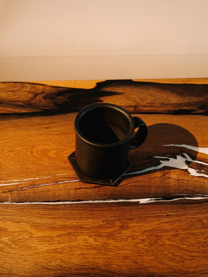 May include: A black ceramic mug with a handle sits on a wooden table with white streaks. The mug is filled with a dark liquid.