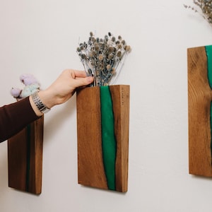 May include: Wooden wall-mounted vase with a vertical green resin stripe. The vase is filled with dried flowers. The wood has a natural grain and is rectangular. The vase is mounted on a white wall.