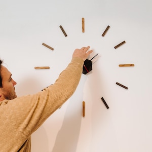 May include: A person adjusts the hands of a black hexagonal clock on a white wall. The clock has wooden hour markers that are arranged in a circle around the clock face.