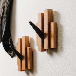 May include: Two wooden wall hooks with a geometric design. The hooks are made of light brown wood with a black, triangular hook extending from the center. The hooks are mounted on a white wall.