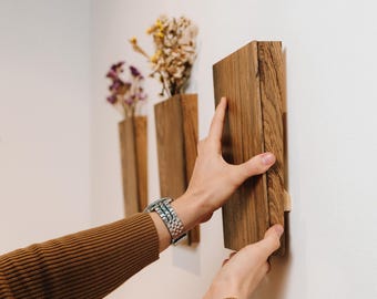 May include: Wooden wall-mounted vases in a row, each holding dried flowers. The vases are rectangular and made of light brown wood. The image shows a person installing one of the vases on a white wall.