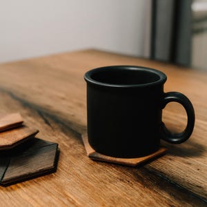 May include: A black ceramic mug sits on a wooden coaster with a hexagonal shape. The coaster is made of bog oak wood and has a natural wood grain. The mug is on a wooden table with a light brown finish.