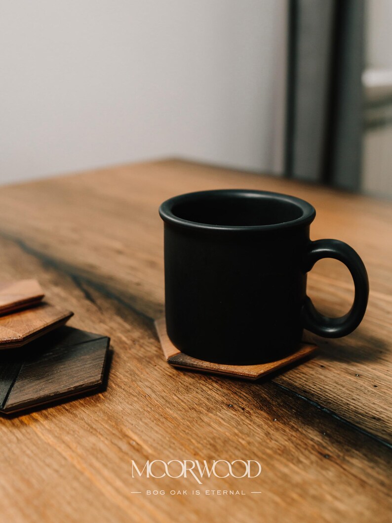 May include: A black ceramic mug sits on a wooden coaster with a hexagonal shape. The coaster is made of bog oak wood and has a natural wood grain. The mug is on a wooden table with a light brown finish.
