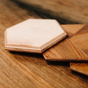 May include: Two wooden coasters, one hexagonal and one with a geometric design, made from bog oak. The coasters are on a wooden surface. The text "MOORWOOD - BOG OAK IS ETERNAL -" is visible below the coasters.