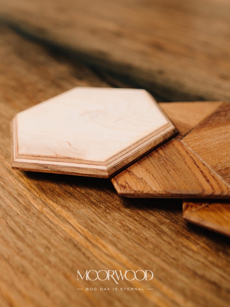 May include: Two wooden coasters, one hexagonal and one with a geometric design, made from bog oak. The coasters are on a wooden surface. The text "MOORWOOD - BOG OAK IS ETERNAL -" is visible below the coasters.