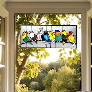 May include: A stained glass window panel featuring a row of colorful birds perched on a branch. The birds are a mix of yellow, blue, green, red, and white. The panel is framed in black metal and has a chain hanging from the top.