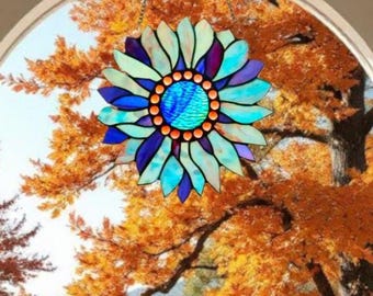 Può includere: Un acchiappasole a forma di girasole in vetro colorato con una combinazione di colori blu, verde acqua e viola. Il fiore è appeso davanti a una finestra con vista sul fogliame autunnale. L'acchiappasole è sospeso da una catena.