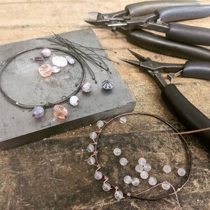 K&ouml;nnte beinhalten: Nahaufnahme von zwei Drahtarmb&auml;ndern, die mit verschiedenen Perlen hergestellt werden. Die Armb&auml;nder liegen auf einer grauen Oberfl&auml;che mit Drahtschneidern und Zangen in der N&auml;he.