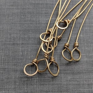 May include: A set of six gold-toned metal jump rings with a variety of shapes and sizes. The rings are arranged in a pile on a gray textured surface.