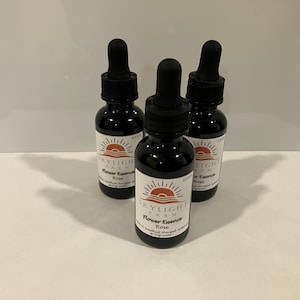 May include: Three small brown glass bottles with dropper tops filled with a clear liquid. Each bottle has a white label with the text "Skylight Farm" and "Flower Essence Rose" printed on it. The label also features a graphic of a sun rising over a field.