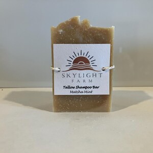 May include: A light brown tallow shampoo bar with a textured surface. A white label with the text "Skylight Farm Tallow Shampoo Bar Matcha Mint" is tied around the bar with twine. The label features a sun and wave logo.