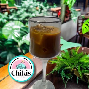 Puede incluir: Un vaso de café helado en un vaso transparente con tallo. El café es de color marrón claro y tiene cubitos de hielo. El vaso está sobre una mesa de madera con una planta verde en el fondo. El logotipo de "Chikis Velas y Jabones" está en primer plano.