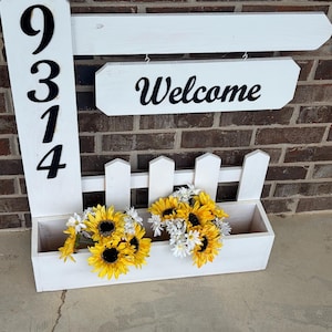 May include: A white wooden welcome sign with the address numbers 9314 in black. The sign has a planter box filled with yellow sunflowers and white daisies. The word "Welcome" is written in black script.