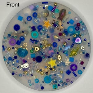 Puede incluir: Un posavasos redondo de resina con un diseño de temática marina. Presenta conchas marinas, estrellas de mar y varias cuentas azules y moradas. El posavasos tiene un acabado transparente y brillante.