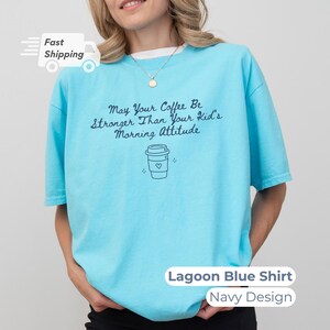 A model wearing a lagoon blue t-shirt with navy blue text reading May Your Coffee Be Stronger Than Your Kid's Morning Attitude and a matching coffee cup graphic.