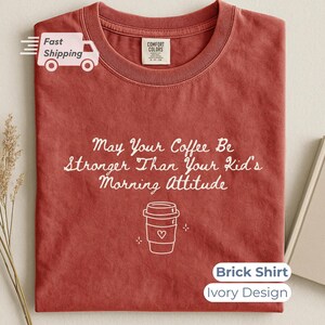 A brick-red t-shirt laid flat with white printed text and a coffee cup design, accompanied by a book and dried flowers on a neutral surface.