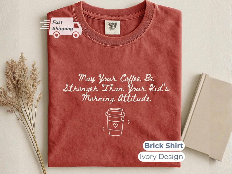 A brick-red t-shirt laid flat with white printed text and a coffee cup design, accompanied by a book and dried flowers on a neutral surface.