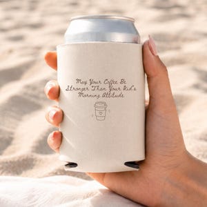 A hand holding a white can cooler printed with the May Your Coffee Be Stronger Than Your Kid's Morning Attitude design against a sandy background.