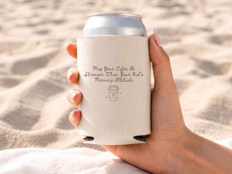 A hand holding a white can cooler printed with the May Your Coffee Be Stronger Than Your Kid's Morning Attitude design against a sandy background.