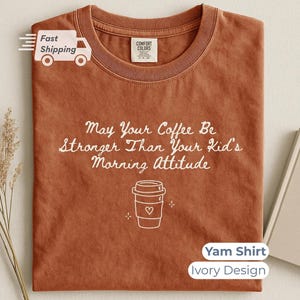 A yam-colored t-shirt with white text reading May Your Coffee Be Stronger Than Your Kid's Morning Attitude and a coffee cup graphic, laid flat with a book and dried plant.