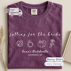 May include: A berry-coloured crew neck t-shirt featuring an ivory graphic design. The text reads falling for the bride, Emma's Bachelorette, and SAVANNAH, GA. Icons include a maple leaf, a diamond ring, a pumpkin, and a champagne bottle.