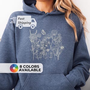Pu&ograve; includere: Una felpa con cappuccio blu con un design floreale beige chiaro. La felpa ha una tasca frontale e un cappuccio. Il testo "8 COLORS AVAILABLE" &egrave; visualizzato in basso a sinistra nell'immagine.