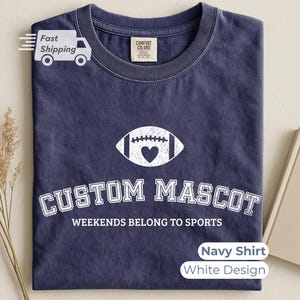 May include: A navy blue crew neck t-shirt featuring a white graphic of a football with a heart in the center. Below the football, the text reads CUSTOM MASCOT in a collegiate font, followed by the text WEEKENDS BELONG TO SPORTS in a sans-serif font.