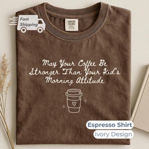 An espresso brown t-shirt with white text and a coffee cup outline, styled flat next to a closed notebook and a small bouquet of dried wheat.