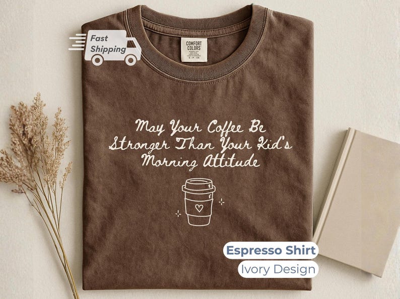 An espresso brown t-shirt with white text and a coffee cup outline, styled flat next to a closed notebook and a small bouquet of dried wheat.