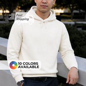 May include: Cream-colored hoodie with a drawstring hood. The image shows a person wearing the hoodie. The text "10 COLORS AVAILABLE" is displayed on the bottom left. The text "Fast Shipping" is displayed on the top left.