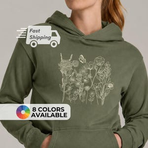 Pu&ograve; includere: Una felpa con cappuccio verde oliva con un design floreale color crema e una farfalla. La felpa ha una tasca a marsupio e un cappuccio con coulisse. Il testo "8 COLORS AVAILABLE" &egrave; visualizzato in basso a sinistra.