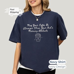 A person wearing a navy blue t-shirt with white lettering that reads May Your Coffee Be Stronger Than Your Kid's Morning Attitude and a coffee cup graphic.