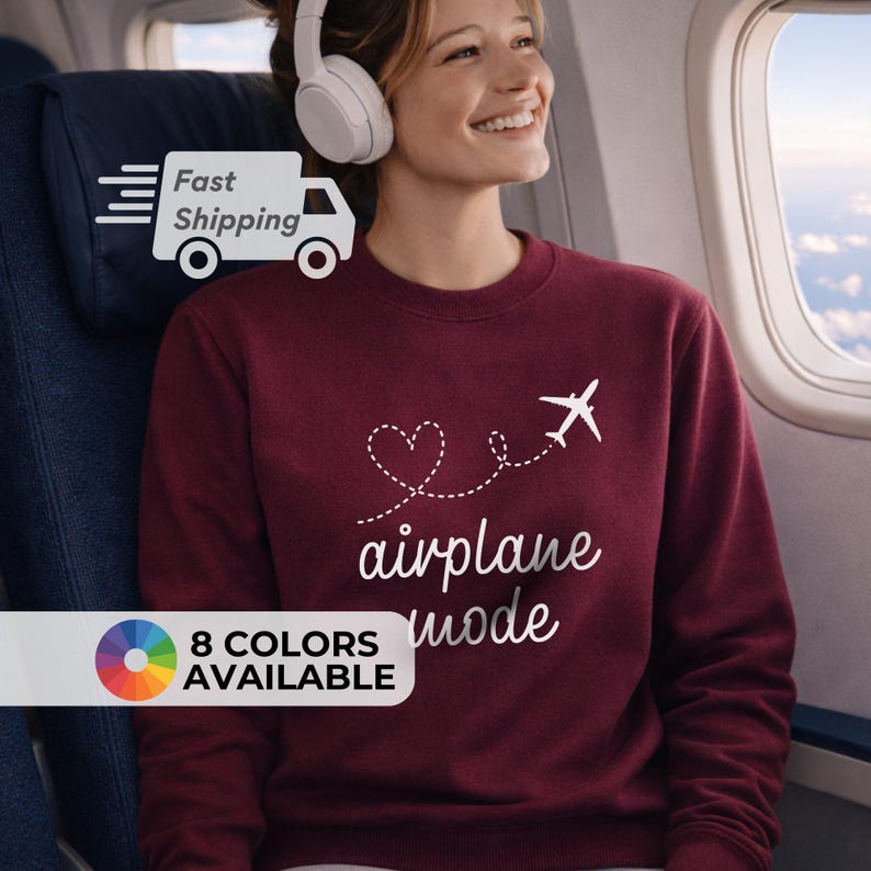 May include: A maroon sweatshirt with the words "airplane mode" in white script, along with a heart and airplane graphic. The sweatshirt is shown on a person wearing headphones on an airplane. The text "8 COLORS AVAILABLE" is also visible.