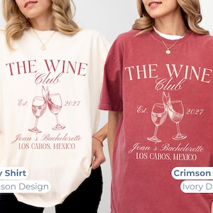 May include: Two t-shirts, one ivory and one crimson, each with a wine-themed graphic. The design includes the text "THE WINE Club Est. 2027" and "Joan's Bachelorette LOS CABOS, MEXICO" with wine glasses. The shirts are short-sleeved.