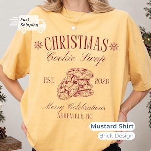 Op de afbeelding: Een mosterdgeel t-shirt met een donkerrode opdruk met de tekst CHRISTMAS Cookie Swap, EST. 2026, Merry Celebrations en ASHEVILLE, NC. Het ontwerp bevat een stapel koekjes met chocoladestukjes en twee sneeuwvlokpictogrammen.