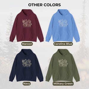 Pu&ograve; includere: Esposizione di quattro felpe con cappuccio in vari colori: bordeaux, blu Carolina, blu navy e verde militare. Ogni felpa presenta un disegno floreale sul davanti e le parole "OTHER COLORS" in alto. I colori sono etichettati sotto.