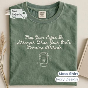 A moss green t-shirt with white text reading May Your Coffee Be Stronger Than Your Kid's Morning Attitude and a coffee cup graphic, laid flat with a closed notebook and dried grass.