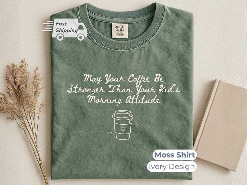 A moss green t-shirt with white text reading May Your Coffee Be Stronger Than Your Kid's Morning Attitude and a coffee cup graphic, laid flat with a closed notebook and dried grass.