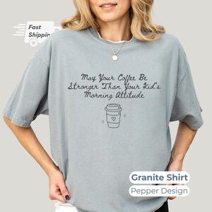 A granite gray t-shirt worn by a smiling model, featuring pepper-colored text and a simple coffee cup graphic.