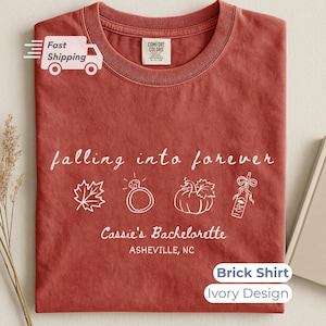 May include: A brick-colored t-shirt featuring an ivory-colored graphic with the text falling into forever, Cassie's Bachelorette, and ASHEVILLE, NC. The design includes line art of a maple leaf, a diamond ring, a pumpkin, and a champagne bottle.