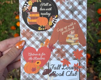 May include: A decorative card with a fall theme, featuring illustrations of pumpkins, leaves, books, and a sweater. The card includes text that reads "Fall in love with reading," "Cozy up with a good book," and "Fall Lovers Book Club."