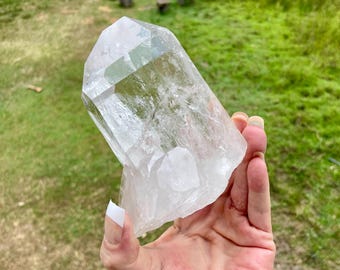 May include: A large, clear, naturally formed quartz crystal point with a multifaceted, translucent structure. The mineral specimen features a sharp, pointed termination and a rugged, irregular base with visible internal inclusions and fractures.