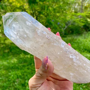 May include: A large, clear quartz crystal with a pointed tip. The crystal is held in a hand against a backdrop of green foliage. The crystal's surface has a textured appearance and a pale, translucent color.