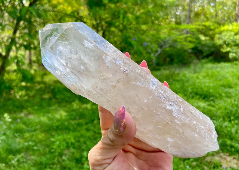 May include: A large, clear quartz crystal with a pointed tip. The crystal is held in a hand against a backdrop of green foliage. The crystal's surface has a textured appearance and a pale, translucent color.