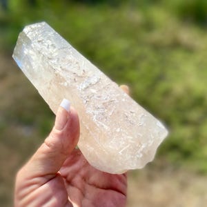 May include: A clear, elongated crystal with a rough, textured surface. The crystal is held in a hand against a blurred green background. The crystal's shape is rectangular with a pointed top.