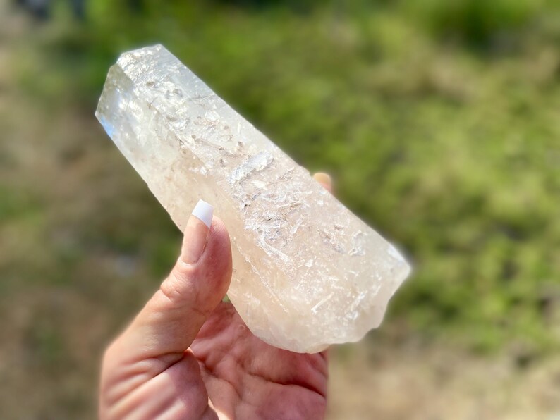 May include: A clear, elongated crystal with a rough, textured surface. The crystal is held in a hand against a blurred green background. The crystal's shape is rectangular with a pointed top.