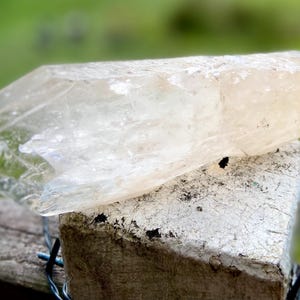 May include: A large, translucent crystal with a light amber hue rests on a weathered, white-painted wooden post. The crystal's surface is textured and reflects light, showcasing its natural form. The background is a soft green.