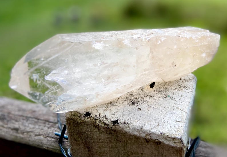 May include: A large, translucent crystal with a light amber hue rests on a weathered, white-painted wooden post. The crystal's surface is textured and reflects light, showcasing its natural form. The background is a soft green.