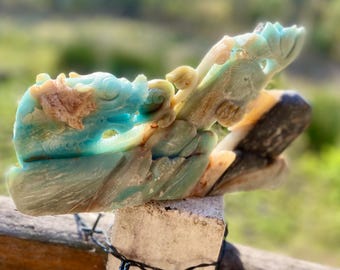 May include: A carved stone dragon sculpture in shades of blue, beige, and brown. The dragon is perched on a stone base, with intricate details visible in its scales and features. The sculpture is set against a blurred green background.
