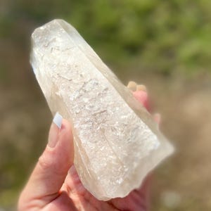 May include: A clear quartz crystal point with a textured surface. The crystal is held in a hand, with a blurred green background. The crystal is a light beige color and has a natural, raw appearance. The crystal is a natural mineral.